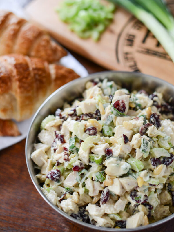 A bowl of cranberry chicken salad with celery and dried cranberries sits on a wooden table. In the background, there are croissants, a cutting board with chopped celery, and green onions.