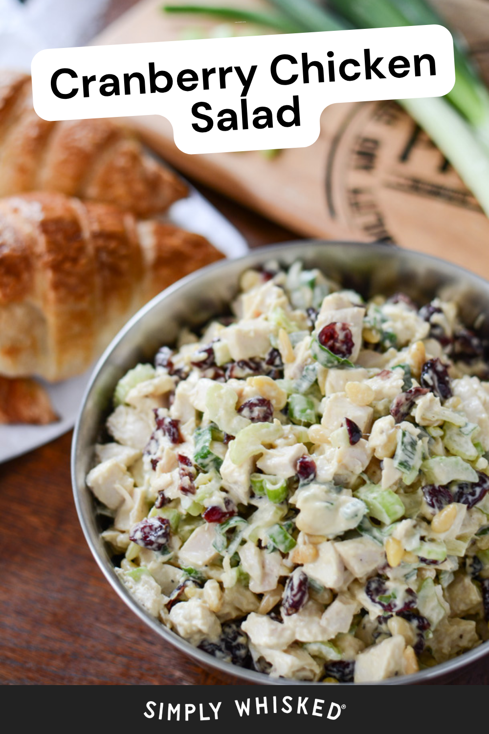 A bowl of cranberry chicken salad sits on a table next to croissants, green onions, and a wooden cutting board, highlighting the fresh ingredients that make this cranberry chicken salad a delicious choice. The text reads, Cranberry Chicken Salad and Simply Whisked.
