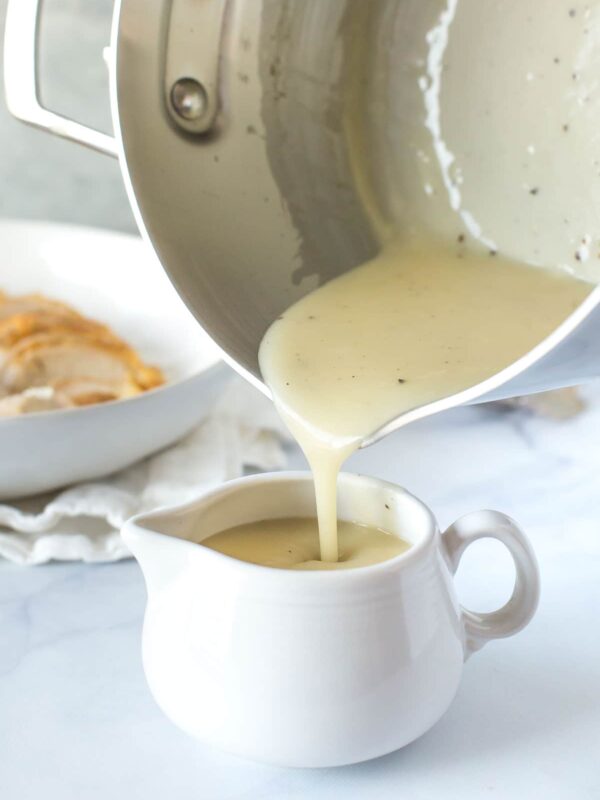chicken velouté sauce, chicken veloute sauce being poured from a saucepan to a white cream pitcher with sliced chicken breast in the background on white marble surface