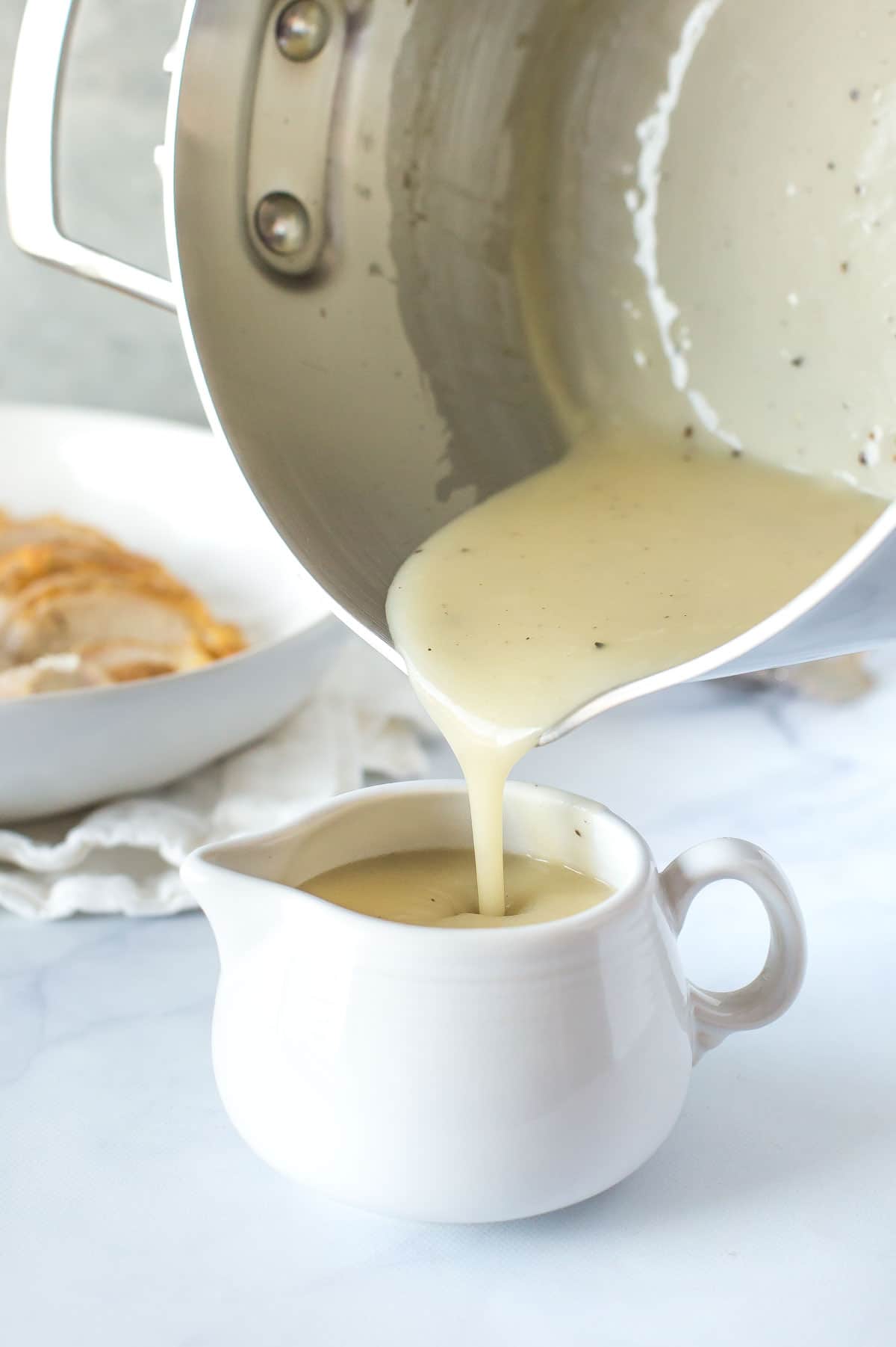 chicken velouté sauce, chicken veloute sauce being poured from a saucepan to a white cream pitcher with sliced chicken breast in the background on white marble surface