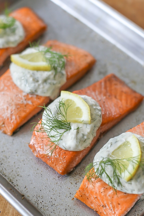 close up side view of cooked salmon with dill sauce, garnished with dill and a lemon wedge on a baking sheet