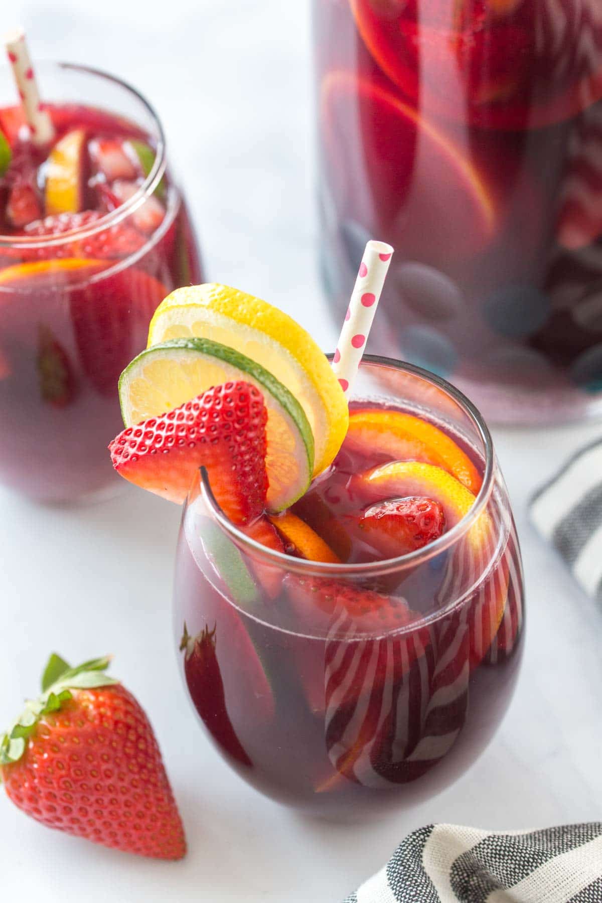 recipe for sangria with red wine in a stemless wine glass garnished with fresh fruit and a polka dot straw, on a bright white background