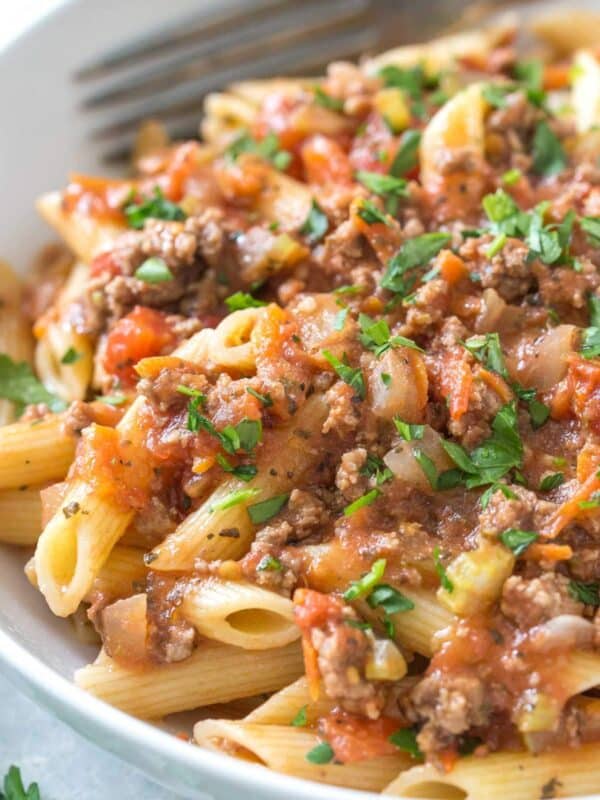 close up of penne topped with bolognese sauce and fresh parsley in a white bowl