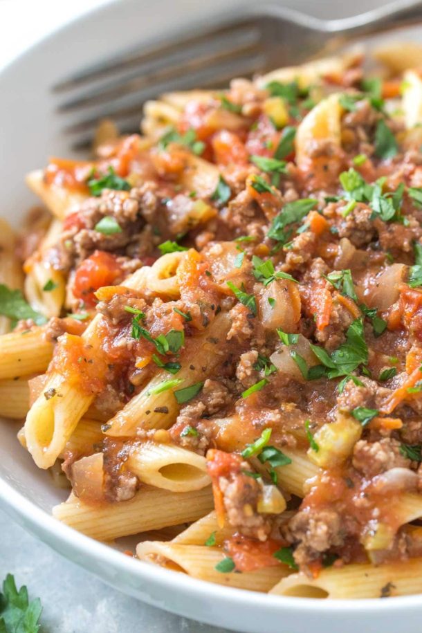 close up of penne topped with bolognese sauce and fresh parsley in a white bowl