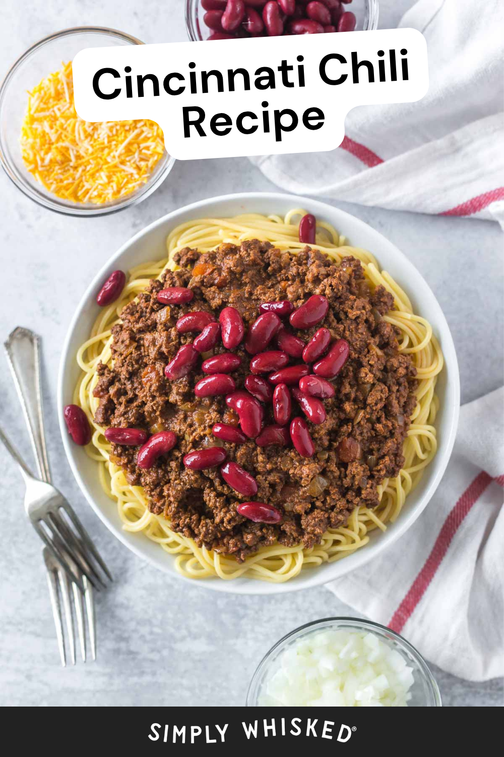 A bowl of spaghetti topped with savory Cincinnati chili and red kidney beans, surrounded by bowls of shredded cheese and chopped onions. A fork, spoon, and striped napkin are beside the plate. Text reads Cincinnati Chili Recipe.