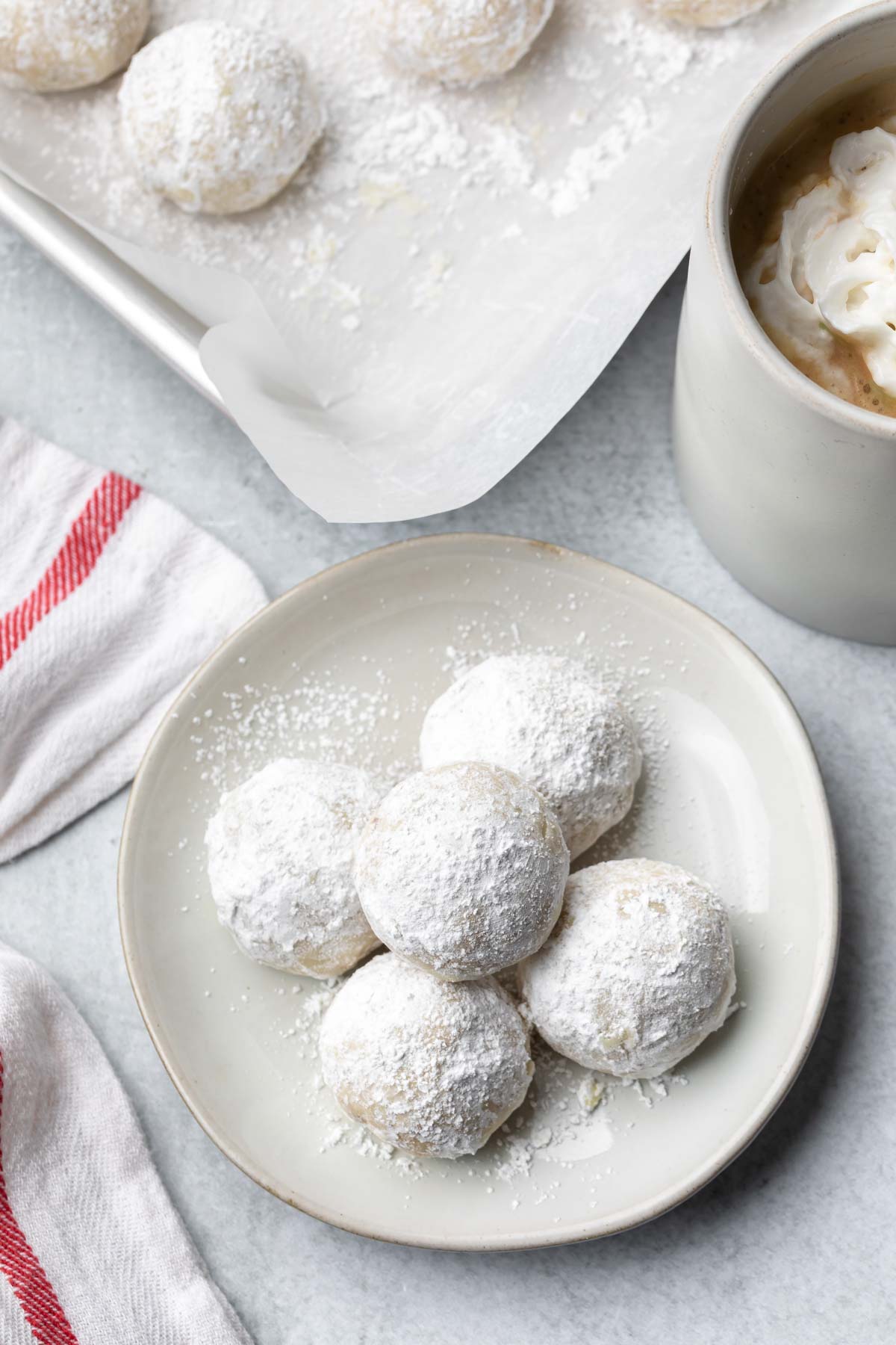overhead image of small plate with 5 pecan snowball cookies on it with a mug of hot chocolate to the side and a baking sheet with more cookies in the background
