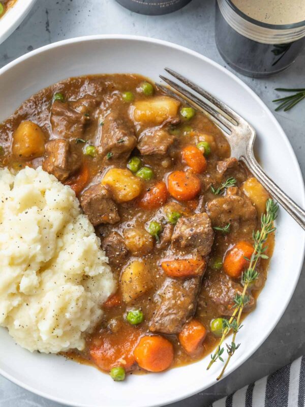 A white bowl filled with Irish beef stew featuring chunks of beef, carrots, potatoes, and peas in rich gravy, served alongside mashed potatoes. A fork sits on the side, and a thyme sprig garnishes the dish.