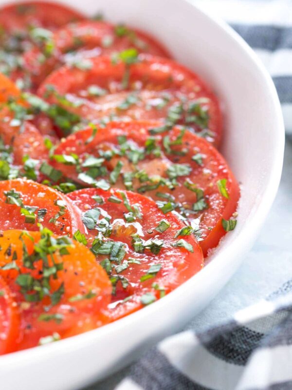Sliced tomato salad topped with herbs and olive oil, in a shallow white bowl with a striped napkin next to it, on a light gray background