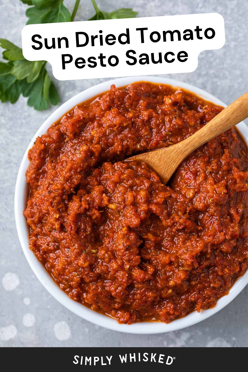 A bowl of chunky sun dried tomato pesto sauce with a wooden spoon, garnished with green herbs, sits invitingly on a gray surface.