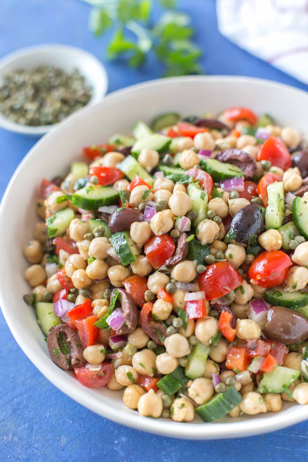 Mediterranean inspired chickpea salad with olives, cucumber, tomatoes, onion, in a white bowl on a blue background sitting next to a small bowl of capers, a parsley sprig and a white and red striped kitchen towel.
