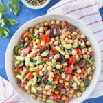 Mediterranean inspired chickpea salad with olives, cucumber, tomatoes, onion, in a white bowl on a blue background sitting on top of a small bowl of capers, a parsley sprig and a white and red striped kitchen towel.
