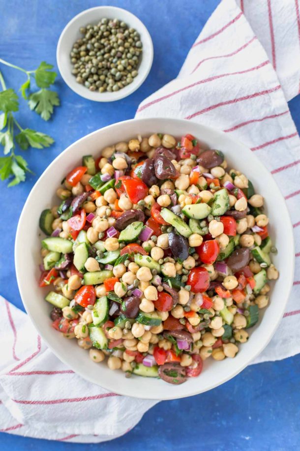 Mediterranean inspired chickpea salad with olives, cucumber, tomatoes, onion, in a white bowl on a blue background sitting on top of a small bowl of capers, a parsley sprig and a white and red striped kitchen towel.