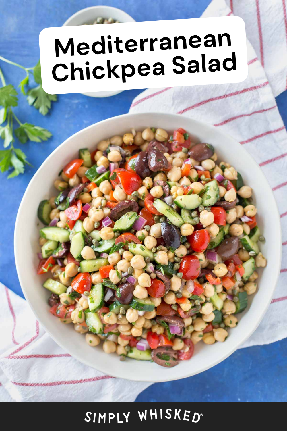 A bowl of vibrant mediterranean chickpea salad with olives, cucumbers, tomatoes, and fresh herbs sits on a blue table next to parsley and a striped towel. Text reads Mediterranean Chickpea Salad.