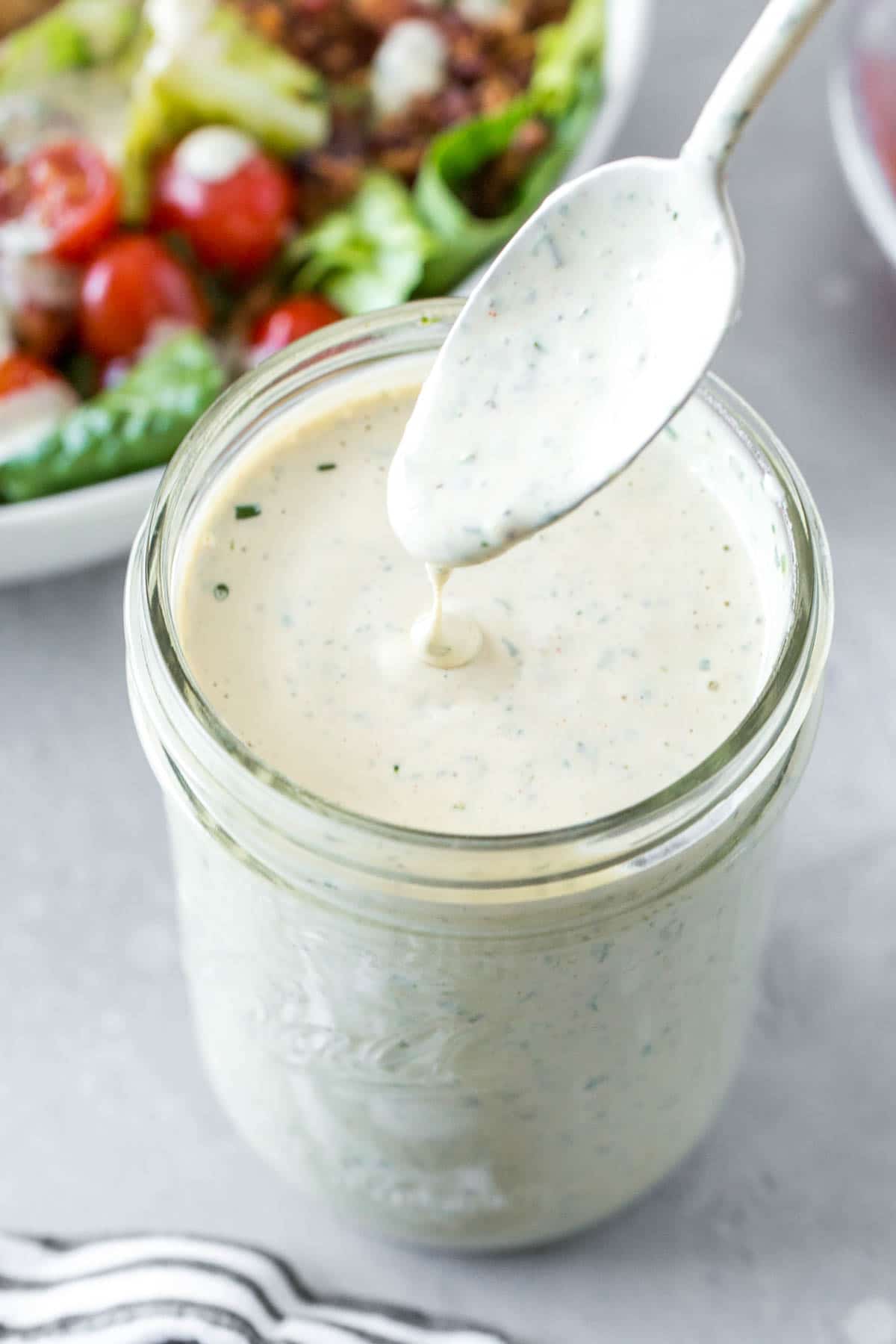 A mason jar of homemade ranch dressing with a spoon removing some and a salad in the background. 