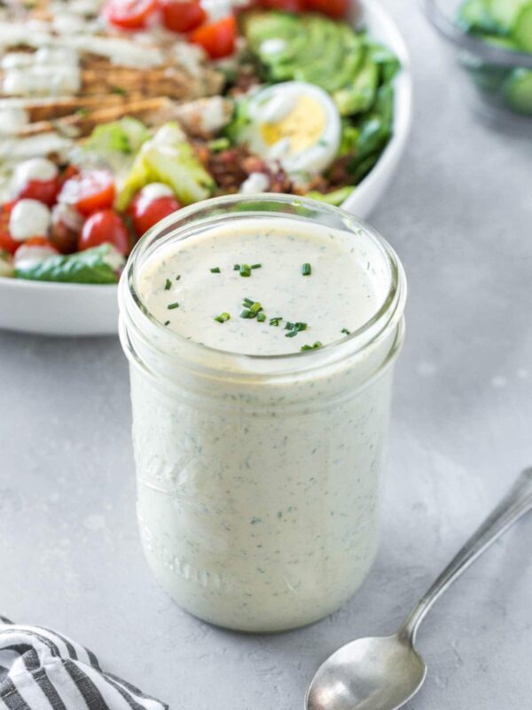 A glass jar filled with creamy homemade ranch dressing, garnished with chopped chives, sits on a gray surface next to a metal spoon. In the background, a salad bowl with assorted vegetables and eggs invites you to try this easy ranch dressing recipe.