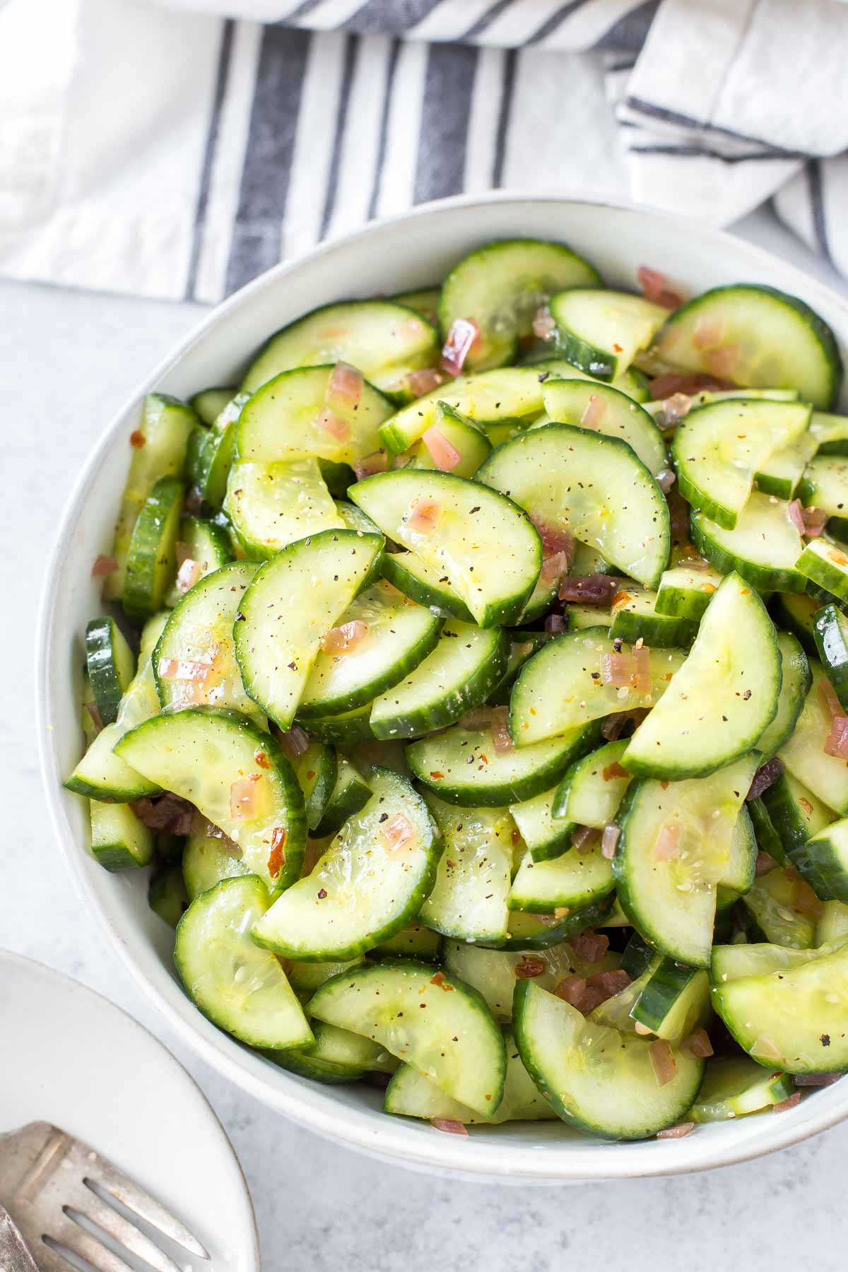 overhead shot of sweet and spicy cucumber salad in a large gray bowl on a light gray background, with a blue and cream striped linen napkin in the above it, a small plate with a vintage fork below it
