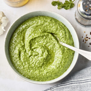 A bowl of vibrant cilantro pesto sauce with a spoon, surrounded by a lemon, a glass bottle of oil, fresh cilantro, red pepper flakes, garlic, and a striped cloth on a light surface.