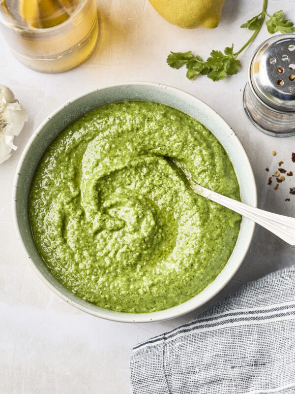 A bowl of vibrant cilantro pesto sauce with a spoon, surrounded by a lemon, a glass bottle of oil, fresh cilantro, red pepper flakes, garlic, and a striped cloth on a light surface.