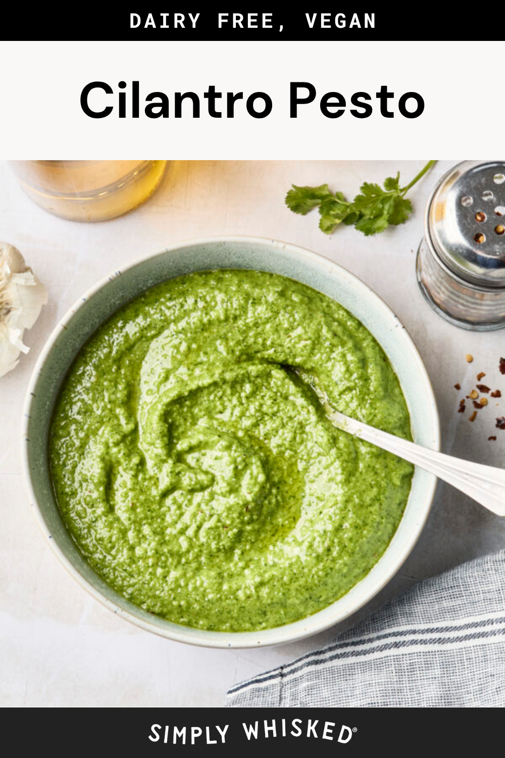 A bowl of vibrant green cilantro pesto with a spoon, surrounded by garlic, fresh cilantro, a shaker, and a striped towel. Text above reads Dairy Free, Vegan Cilantro Pesto—highlighting this flavorful cilantro pesto as the perfect plant-based spread or dip.