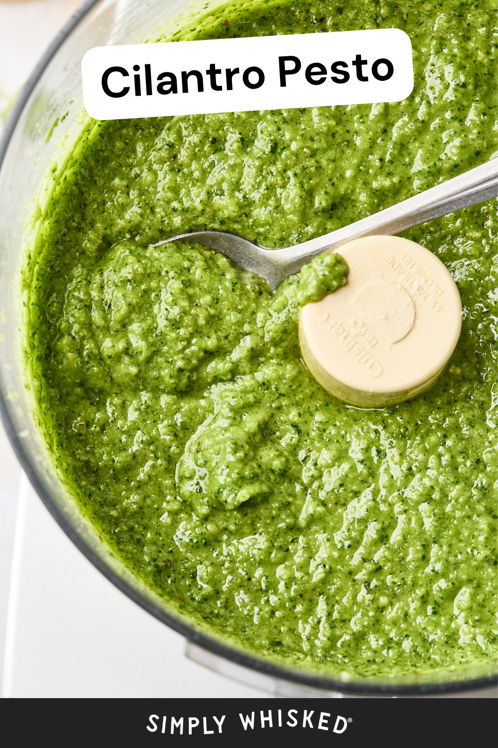 A food processor bowl filled with vibrant cilantro pesto, with a spoon resting inside. Text at the top reads Cilantro Pesto and Simply Whisked appears at the bottom.
