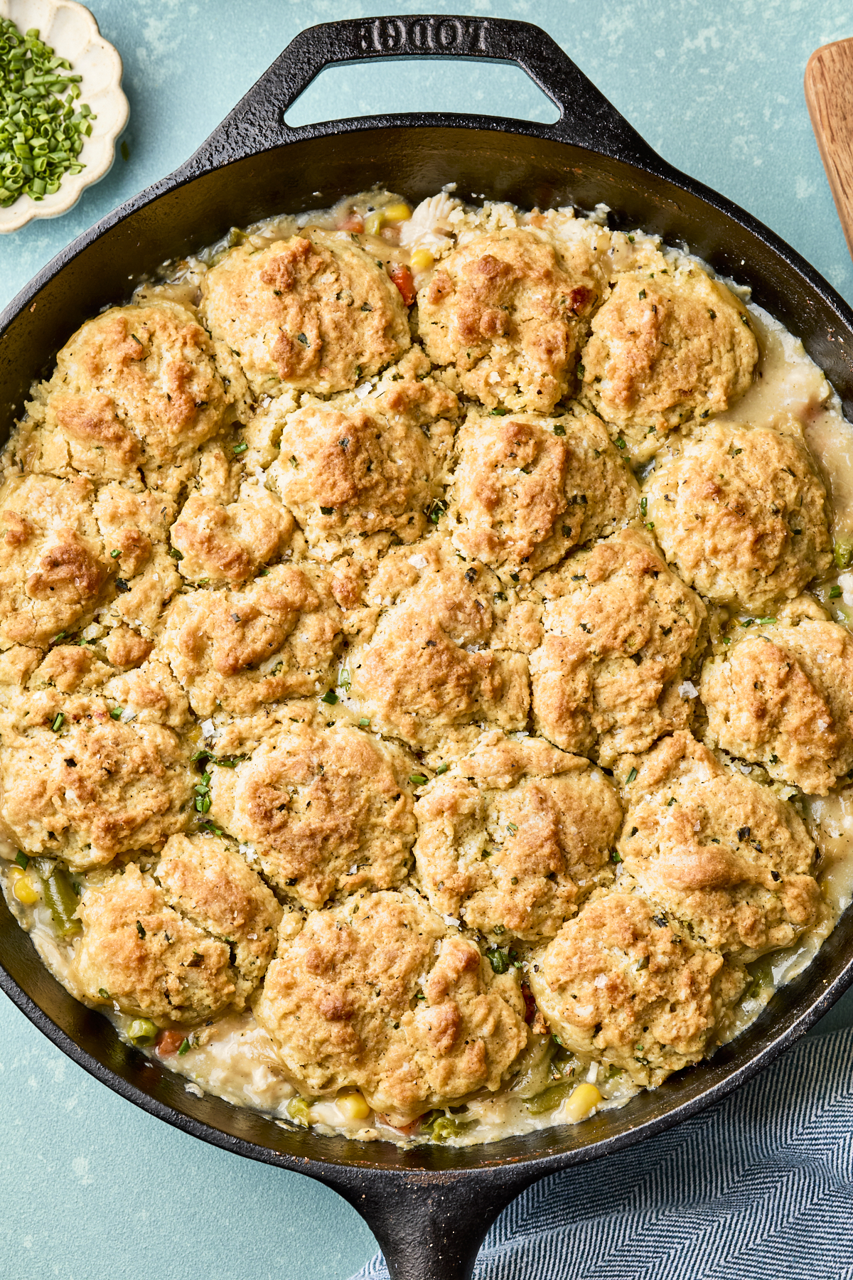A cast iron skillet filled with easy chicken pot pie with biscuits, topped with golden-brown rounds, sits on a blue surface next to a bowl of chopped chives.