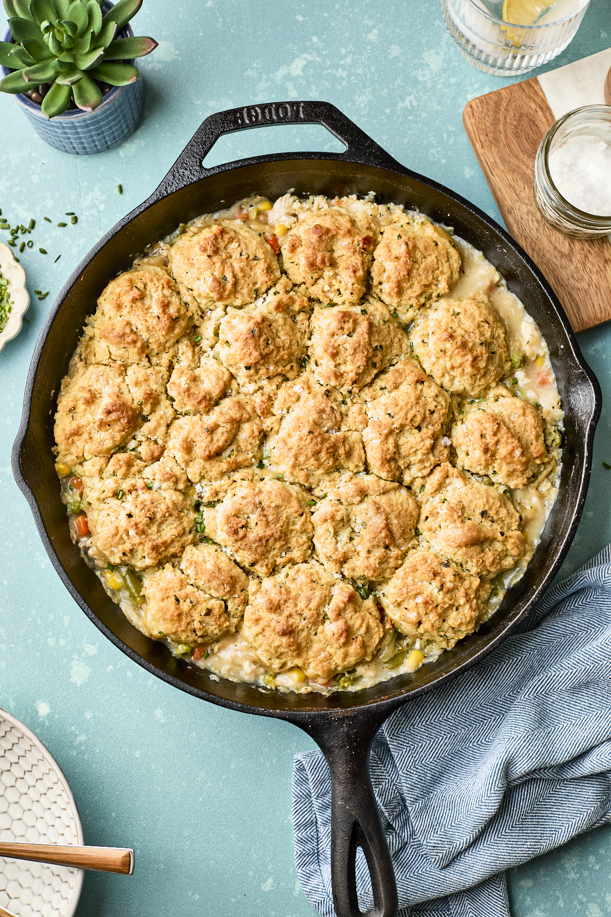 A cast-iron skillet filled with easy chicken pot pie with biscuits, featuring golden, baked biscuits atop a creamy vegetable filling. The skillet rests on a blue background beside a napkin, plates, and a glass of water.