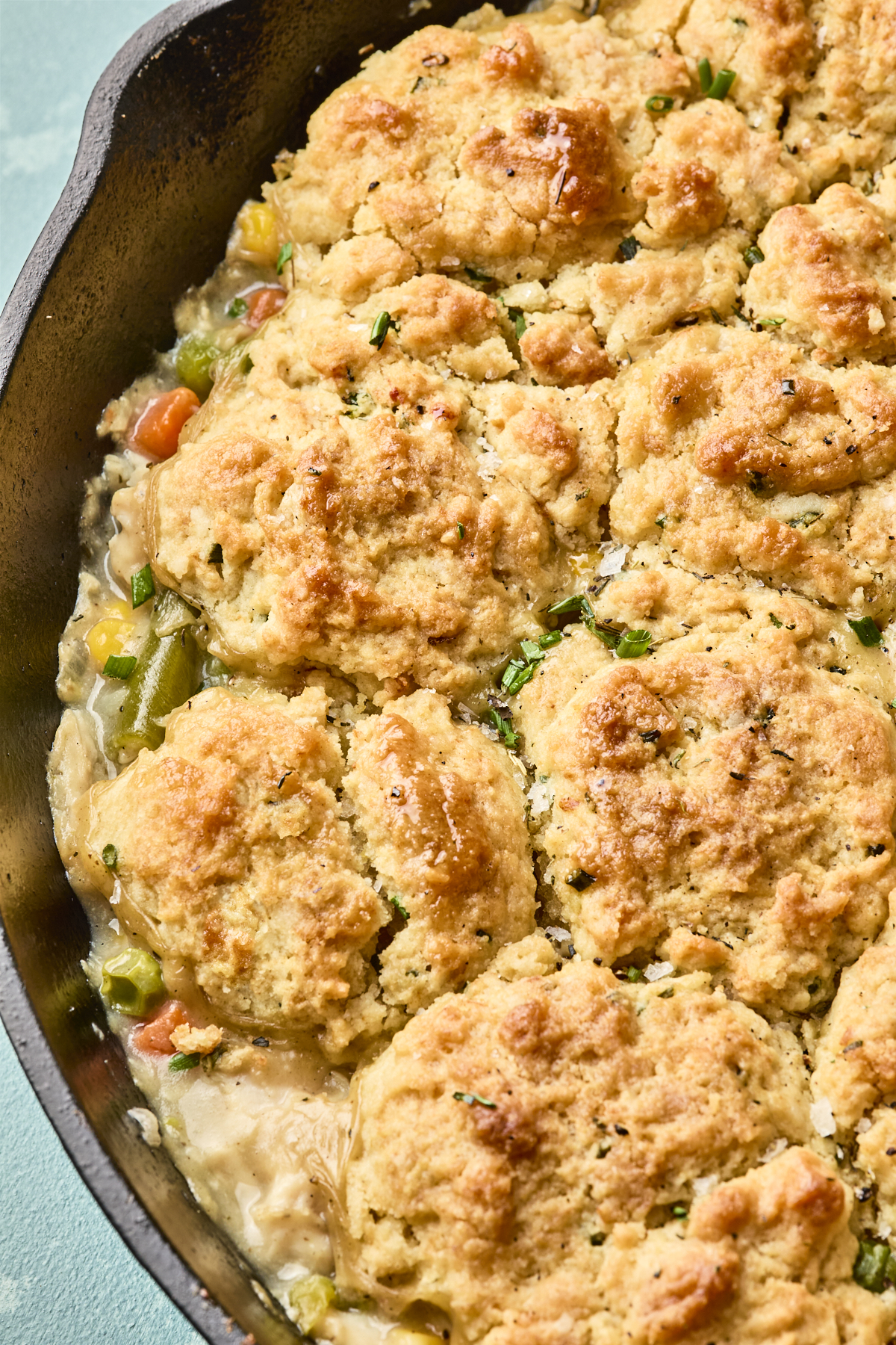 A close-up of an easy chicken pot pie with biscuits in a cast iron skillet, showing golden, flaky biscuit topping. The browned biscuits are slightly crispy, perfectly covering the creamy chicken and vegetable filling below.