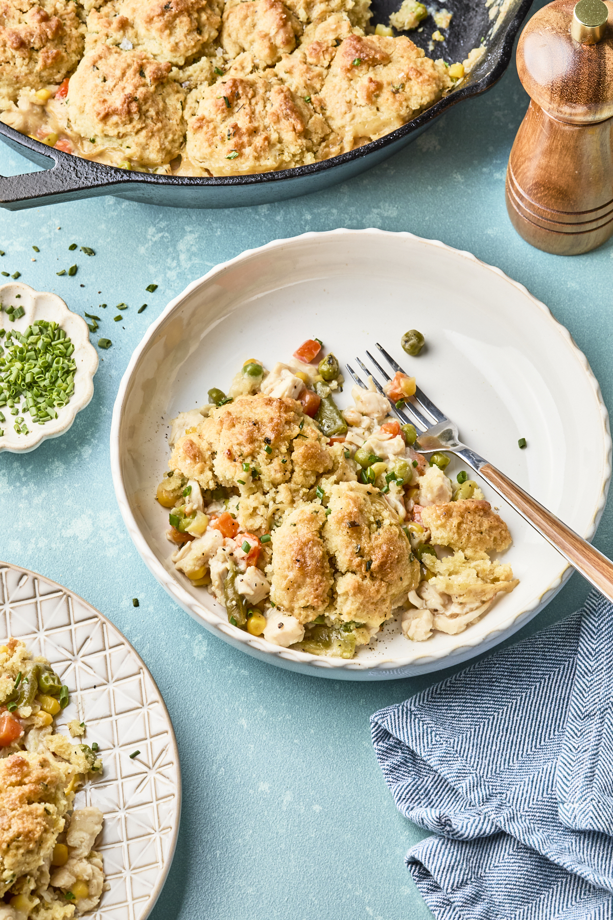 A plate of easy chicken pot pie with biscuits sits on a table next to a skillet of the same dish. Vegetables and chicken are visible, with a fork, napkin, and a small dish of chopped herbs beside the plate.