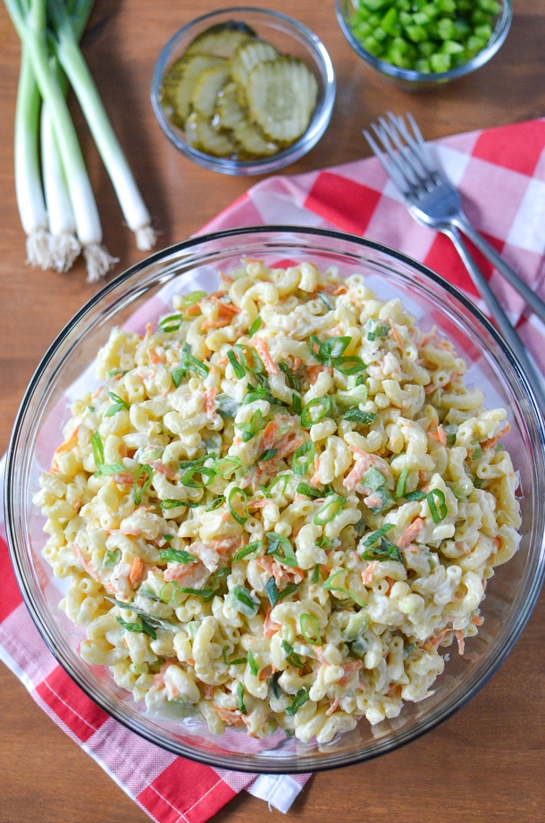 overhead shot of shrimp macaroni salad recipe in a large glass bowl on a red and white gingham napkin with forks, green onions, a bowl of pickles and a bowl of chopped green bell peppers on a wooden board