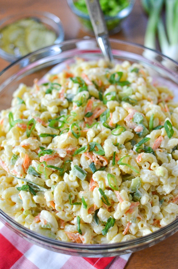 shrimp macaroni salad recipe in a large glass bowl on a red and white gingham napkin, green onions, a bowl of pickles and a bowl of chopped green bell peppers on a wooden board