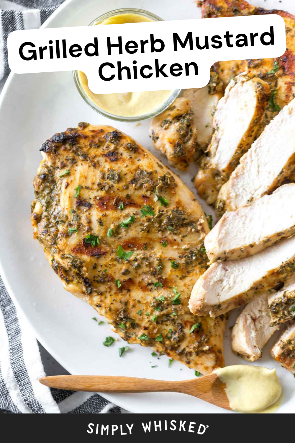 Sliced grilled herb mustard chicken on a white plate with a small bowl of mustard sauce and a wooden spoon, garnished with chopped herbs, makes for a mouthwatering presentation that highlights the rich flavors of classic mustard chicken.