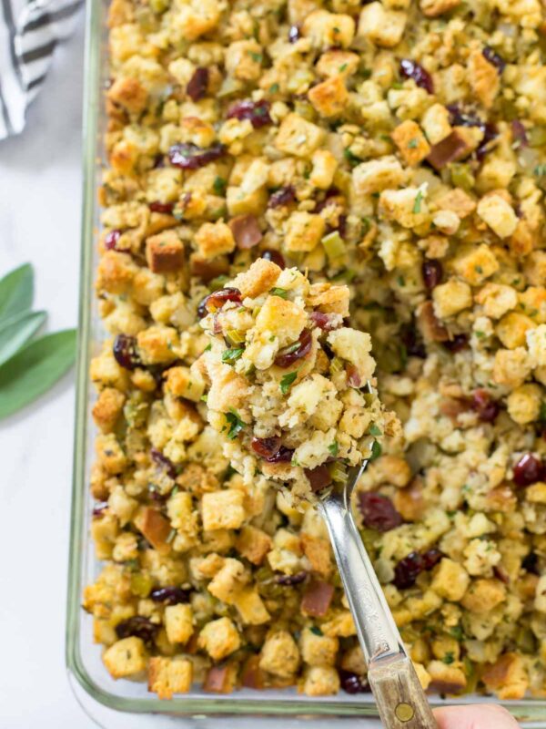 A hand holds a serving spoon filled with golden-brown homemade stuffing, featuring cubed bread, herbs, celery, and dried cranberries, above a glass baking dish full of the same delicious stuffing.