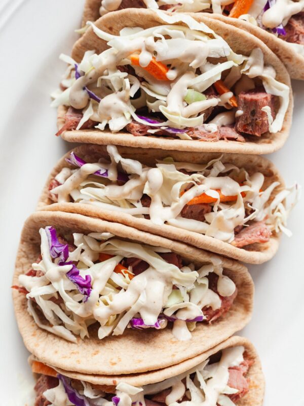 Four soft tacos filled with flavorful corned beef and cabbage, topped with coleslaw of purple and orange cabbage, then drizzled with creamy white sauce, all arranged in a row on a white plate.