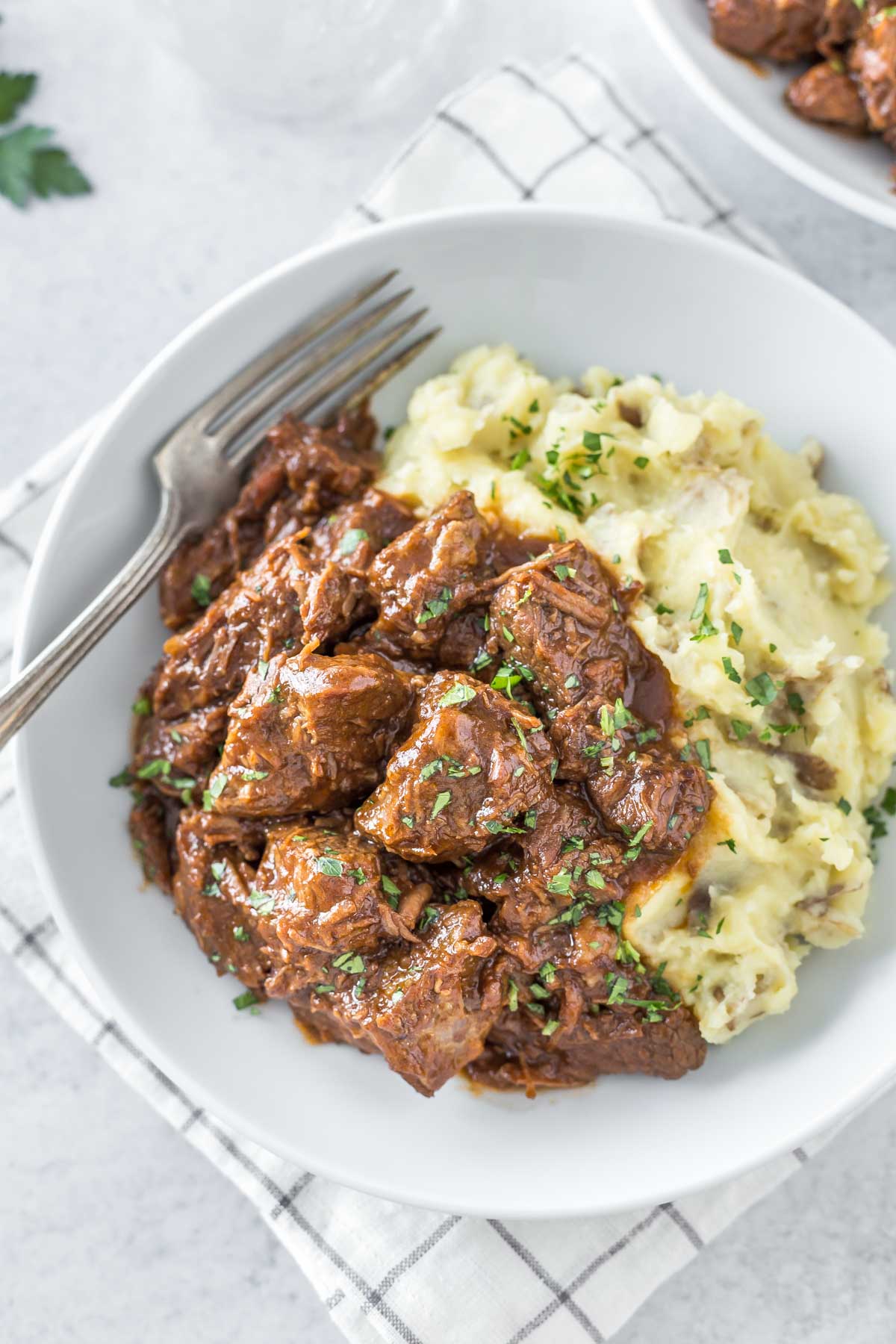 overhead shot of Guinness braised beef in a bowl with mashed potatoes and a vintage fork, on a gray background with a second bowl to the side and a gray and white grid patterned napkin and a sprig of fresh parsley