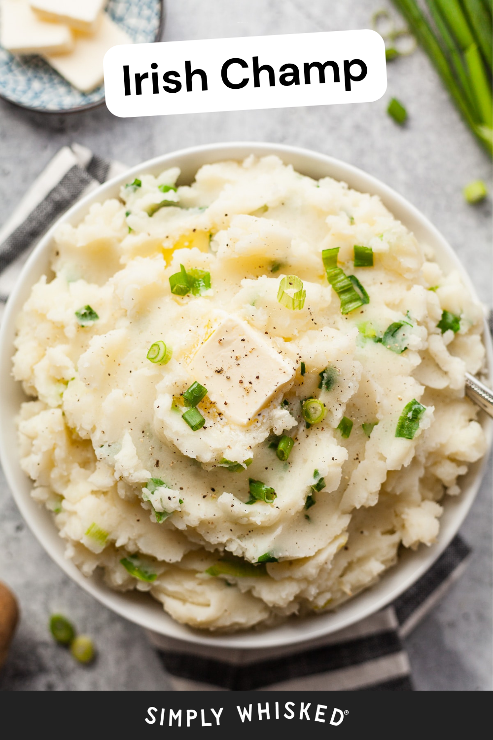A bowl of creamy Irish Champ mashed potatoes, topped with butter, black pepper, and chopped green onions, with a fork and some sliced scallions nearby. This classic Irish Champ dish makes the perfect comforting side for any meal.