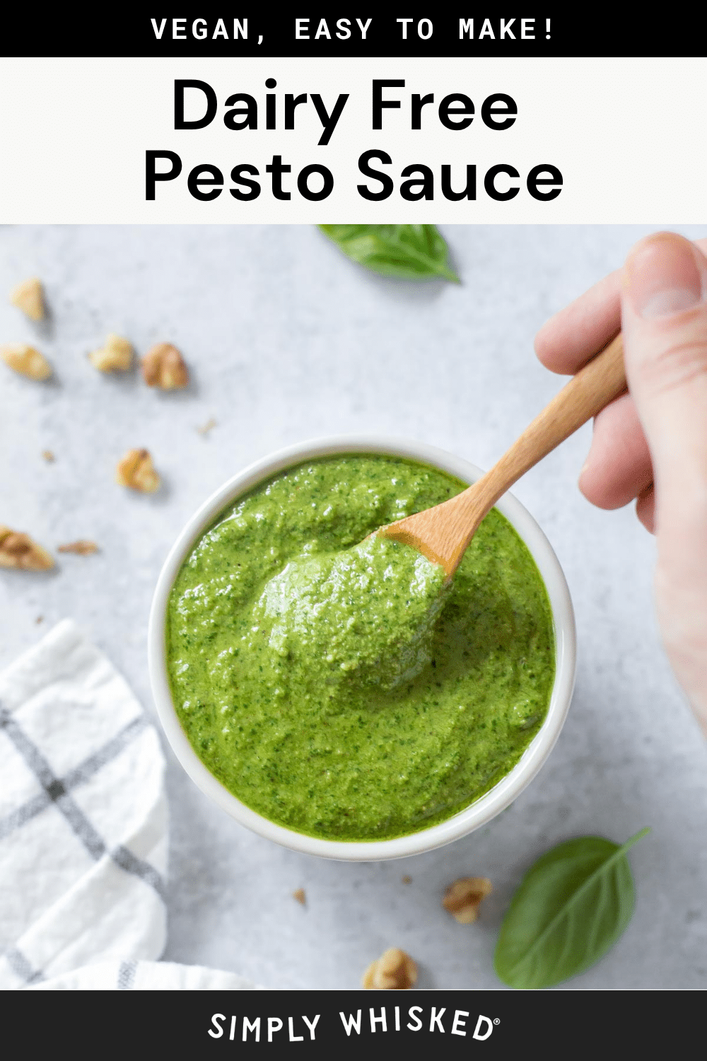 A hand holds a wooden spoon scooping vibrant green Dairy Free Pesto Sauce from a bowl. Walnut pieces and basil leaves are scattered on a light surface near a striped cloth. Text at the top reads, Vegan, Easy to Make! Dairy Free Pesto Sauce.