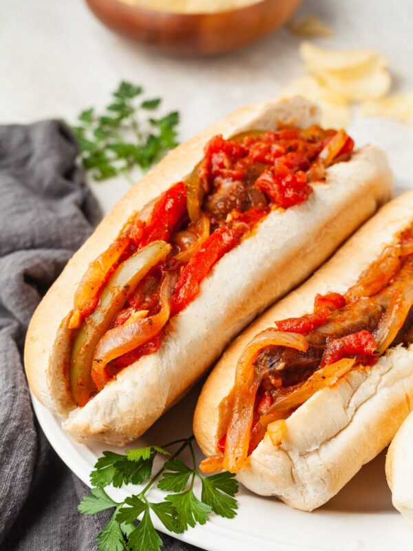 Two hoagie rolls filled with sautéed onions, bell peppers, tomatoes, and savory sausage make up these classic sausage and pepper sandwiches. Garnished with fresh parsley on a white plate, with a gray napkin and bowl of chips in the background.