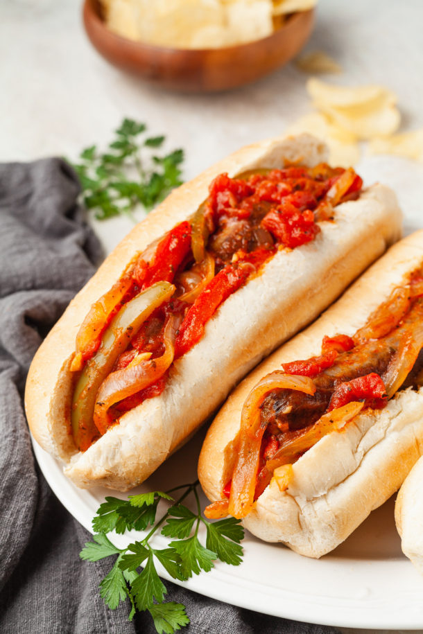 Two hoagie rolls filled with saut&eacute;ed onions, bell peppers, tomatoes, and savory sausage make up these classic sausage and pepper sandwiches. Garnished with fresh parsley on a white plate, with a gray napkin and bowl of chips in the background.