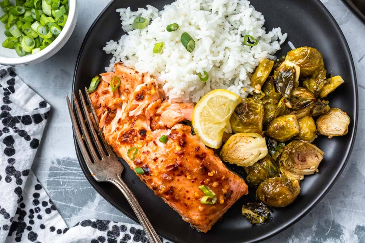overhead image of sweet chili salmon on a black plate with brussels sprouts and riec