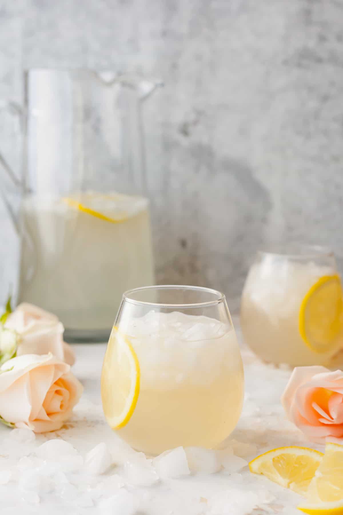 Rose water lemonade in a stemless wine glass with crushed ice and a lemon slice with pale pink roses and more lemon slices scattered around it. A tall glass pitcher of the lemonade in the background.