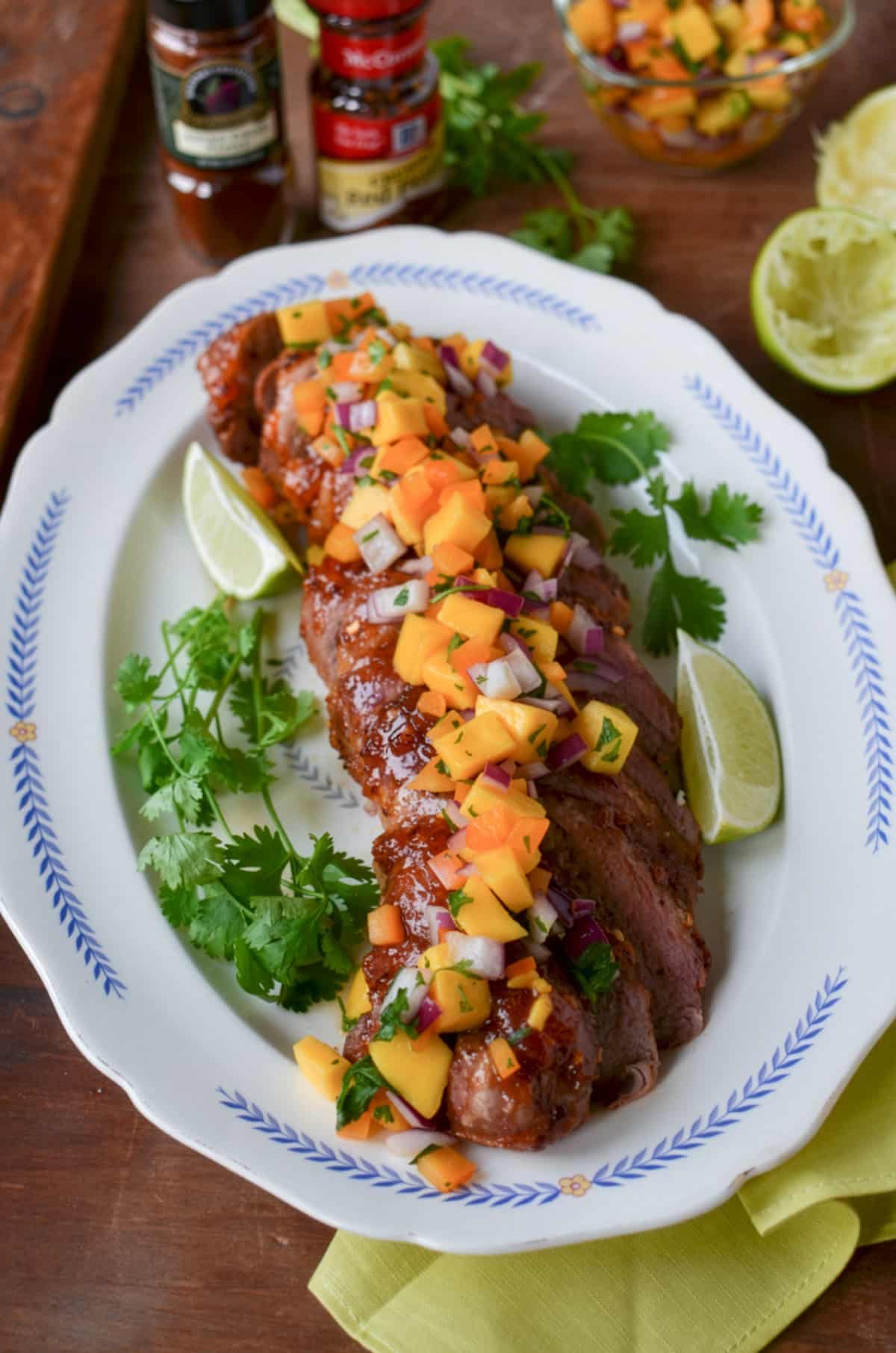 Chamoy glazed pork tenderloin, sliced and topped with colorful mango salsa, is garnished with lime wedges and fresh cilantro. Served on a white oval platter with a blue border, salsa ingredients are visible in the background.