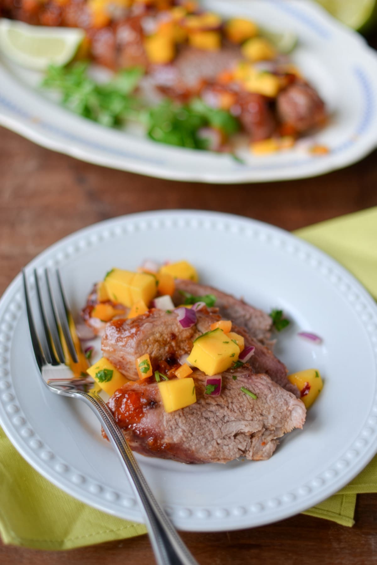 Three slices of chamoy glazed pork tenderloin topped with mango salsa and garnished with herbs are served on a white plate with a fork, set on a yellow napkin. More pork with salsa is visible on a platter in the background.
