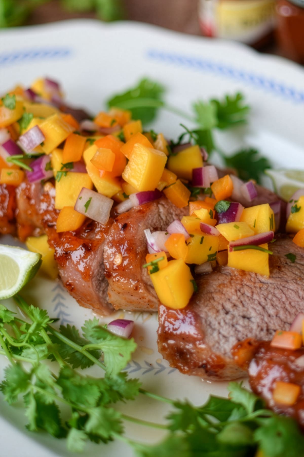 Chamoy glazed pork tenderloin, sliced and topped with a fresh mango salsa of diced mango, red onion, and herbs, served on a white plate with cilantro and lime wedges.