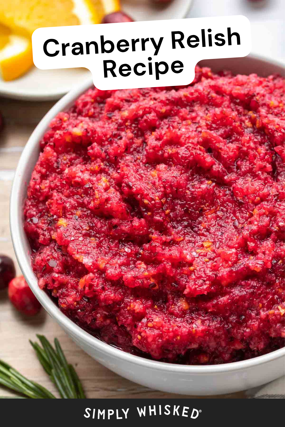 A close-up of a white bowl filled with bright red cranberry relish sits on a table. There are orange slices and herbs in the background, complementing the vibrant cranberry relish. Text on the image reads Cranberry Relish Recipe and Simply Whisked.