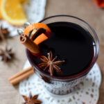 A glass of vin chaud garnished with an orange peel, cinnamon stick, and star anise sits on a lace coaster. Slices of orange and more spices are scattered on the wooden table nearby.