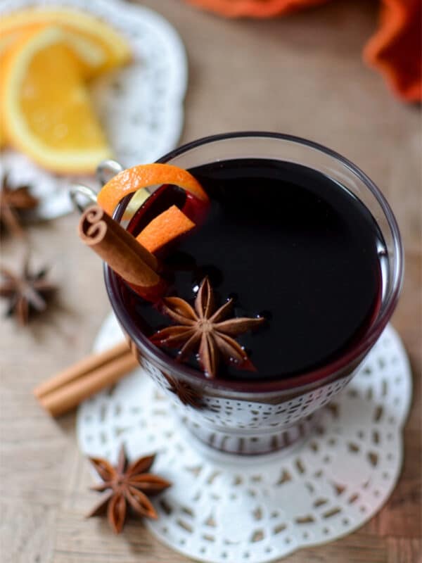 A glass of vin chaud garnished with an orange peel, cinnamon stick, and star anise sits on a lace coaster. Slices of orange and more spices are scattered on the wooden table nearby.