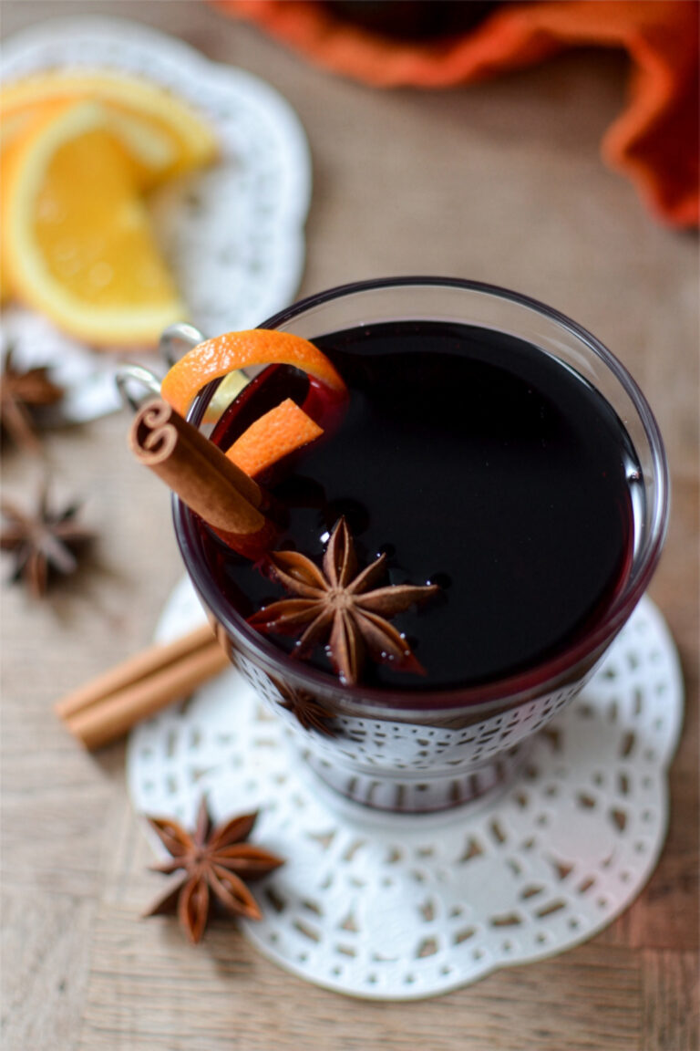 A glass of vin chaud garnished with an orange peel, cinnamon stick, and star anise sits on a lace coaster. Slices of orange and more spices are scattered on the wooden table nearby.
