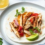 A tortilla topped with sliced grilled chicken marinated in fajita marinade, sautéed onions, red and yellow bell peppers, tomato, cilantro, and a lime wedge on a plate. A drink and another plate of food are partially visible in the background.