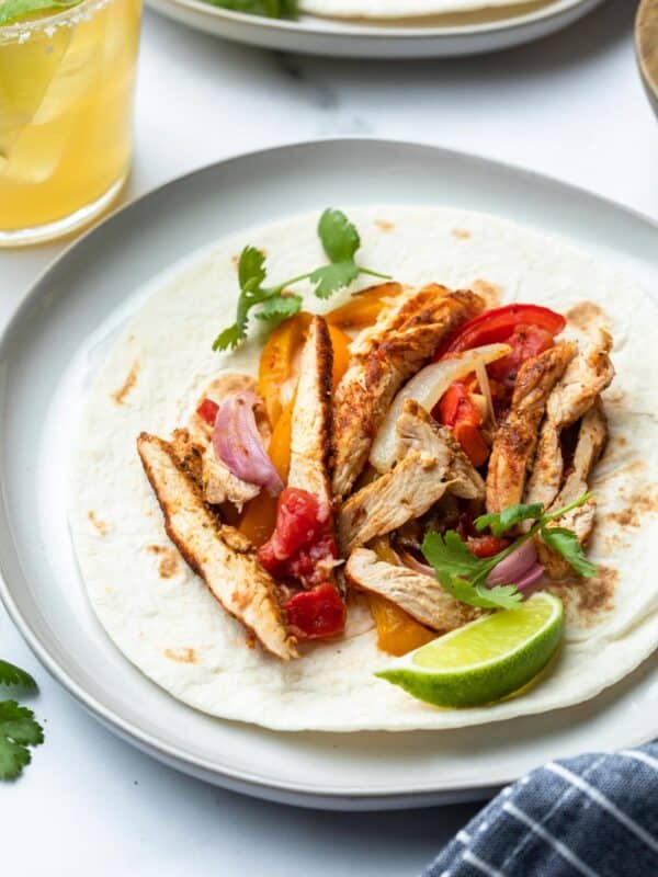 A tortilla topped with sliced grilled chicken marinated in fajita marinade, sautéed onions, red and yellow bell peppers, tomato, cilantro, and a lime wedge on a plate. A drink and another plate of food are partially visible in the background.