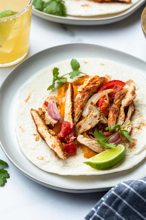 A tortilla topped with sliced grilled chicken marinated in fajita marinade, saut&eacute;ed onions, red and yellow bell peppers, tomato, cilantro, and a lime wedge on a plate. A drink and another plate of food are partially visible in the background.